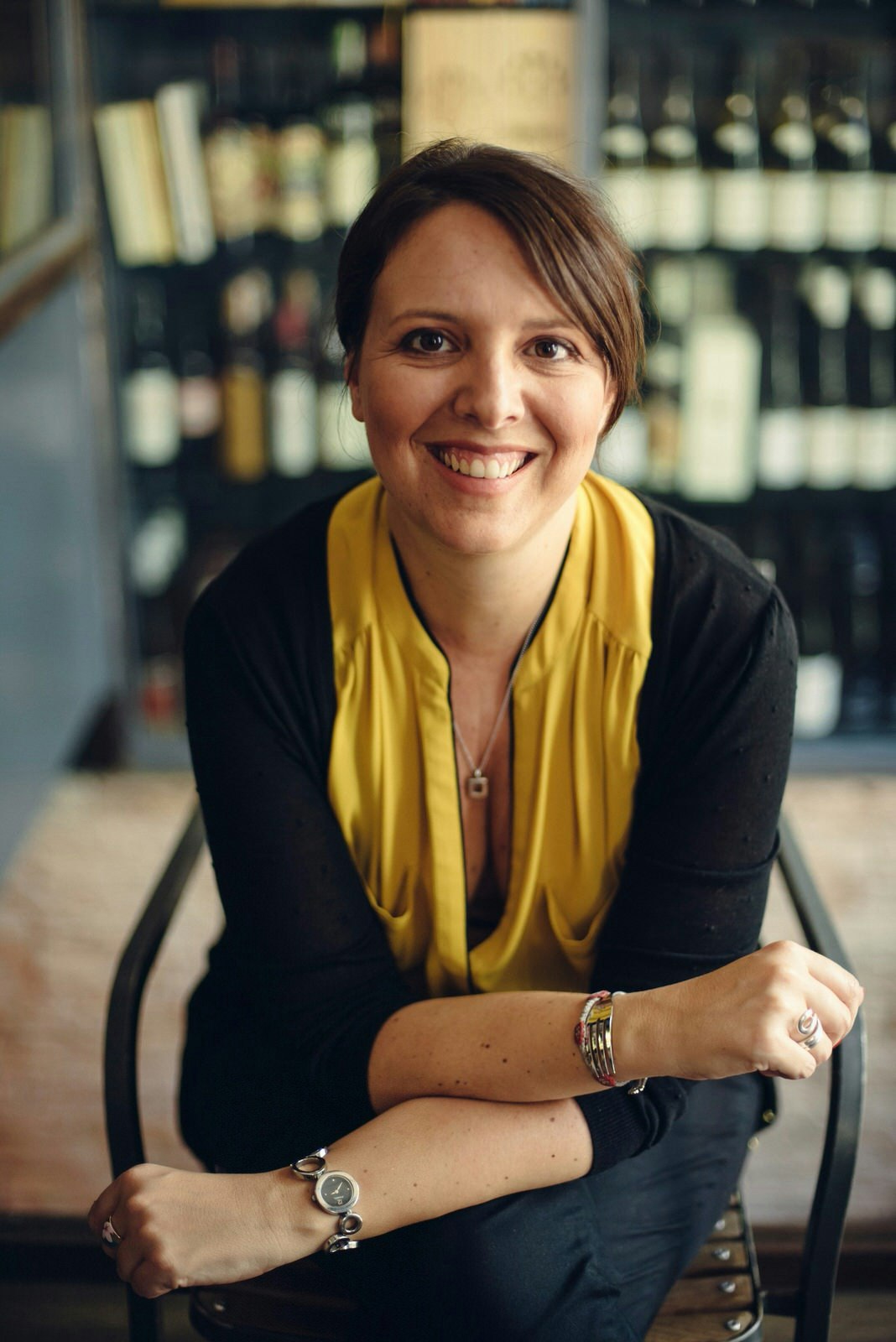 A woman smiling into the camera wearing a yellow scarf and black top. There is wine stacked from floor to ceiling in the background.